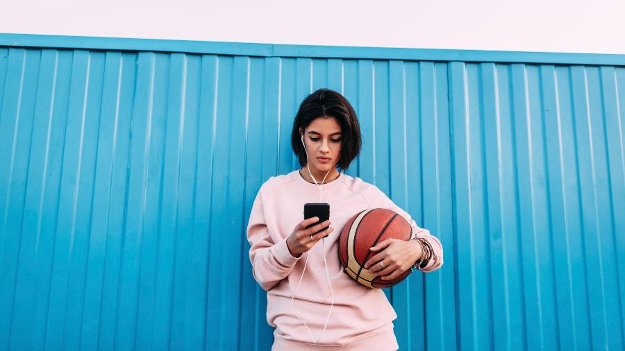 Mulher usa fones de ouvido no celular e segura bola de basquete - Getty Images/Westend61