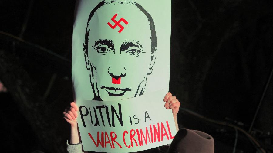 Homem usa cartaz com caricatura de Hitler em foto do presidente russo, Vladimir Putin, em frente à embaixada da Rússia em Vilnius, Lituânia - PETRAS MALUKAS / AFP