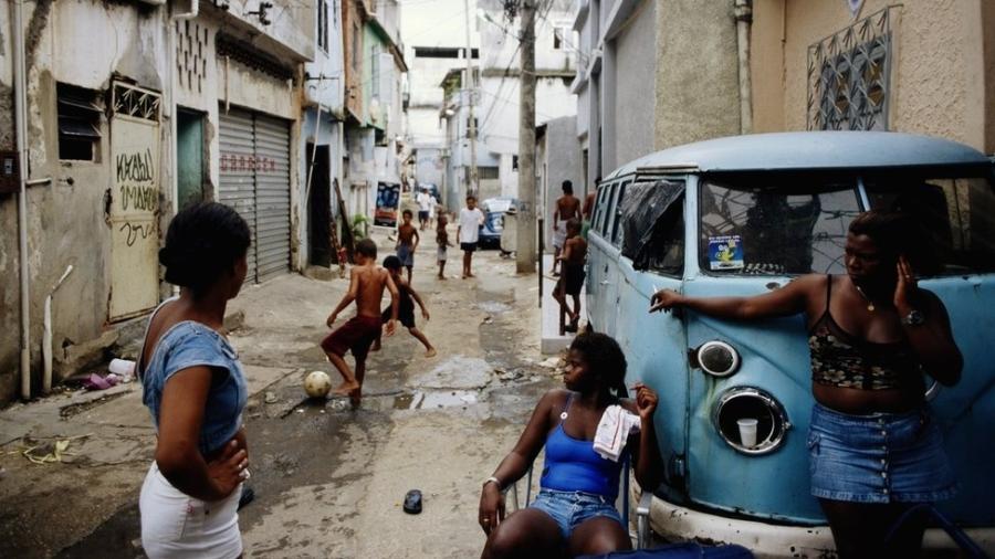 Mulheres conversam apoiadas em uma caminhonete enquanto crianças brincam ao fundo na favela do Jacarezinho, nos anos 1990 - Getty Images/BBC