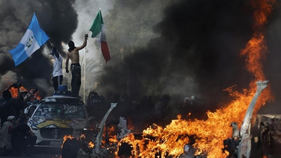 8.jun.2025 - Manifestantes balançam as bandeiras da Guatemala e do México enquanto um carro pega fogo durante uma manifestação contra as políticas migratórias do governo de Donald Trump - MARIO TAMA / GETTY IMAGES NORTH AMERICA / Getty Images via AFP