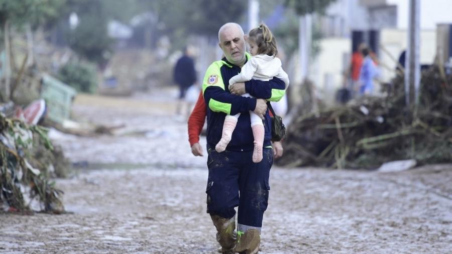 30.out.2024 - Um membro da Proteção Civil carrega uma criança em uma rua coberta de lama em uma área inundada em Picanya, perto de Valência