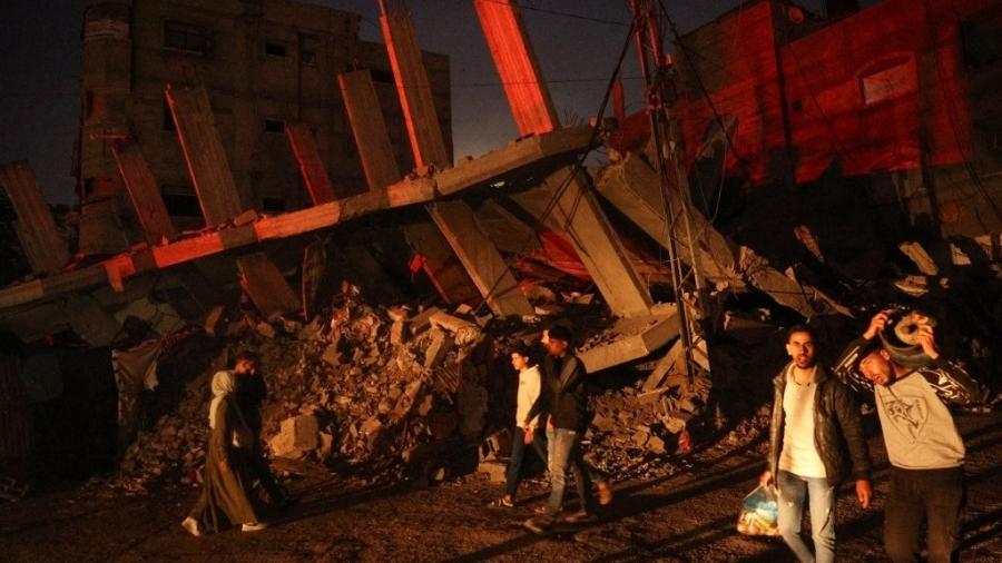 Palestinos passam por um prédio destruído no campo de refugiados de al-Bureij, no centro da Faixa de Gaza, durante a noite de 15 de novembro de 2024