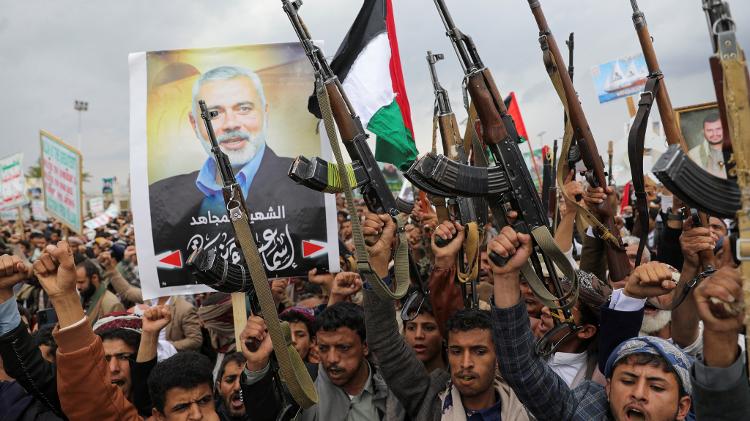 Manifestantes, principalmente apoiadores Houthi, seguram armas de fogo ao lado de um pôster de Ismail Haniyeh no Iêmen