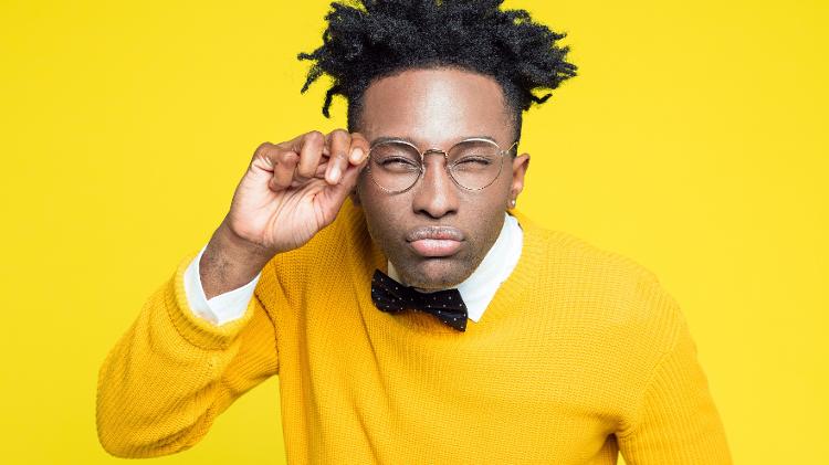 Jovem negro usando blusa amarela e usando óculos fazendo cara de desconfiado, atento, prestando atenção - Getty Images/iStockphoto - Getty Images/iStockphoto