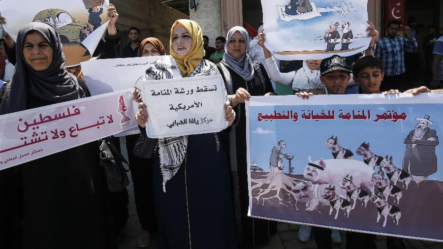 Manifestantes palestinos gritam slogans por trás de um cartaz com os rostos de líderes árabes sobre corpos de cães e porcos e um slogan em árabe: "A conferência de traição e normalização em Manama" durante um protesto contra uma conferência econômica do Oriente Médio patrocinada pelos EUA no Bahrein - Said Khatib/AFP