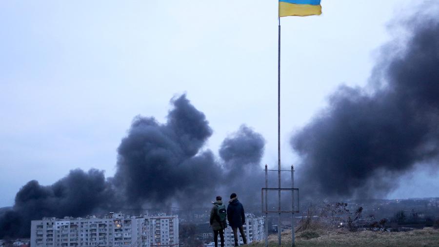 26.mar.2022 - Pessoas em pé perto de uma bandeira nacional ucraniana observam a fumaça escura subir após um ataque aéreo na cidade de Lviv, no oeste da Ucrânia - 26.mar.2022 - Aleksey Filipov/AFP