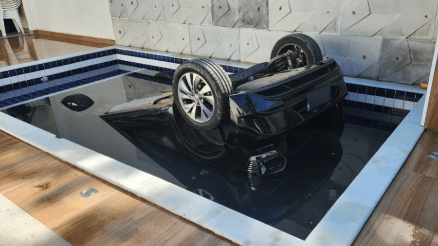 Uma foto mostra o carro preto com as rodas para cima após cair na piscina no Guarujá