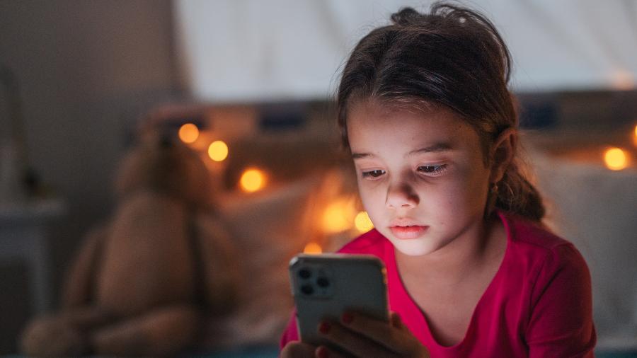 Criança sentada em uma cama, concentrada, olhando para a tela de um celular em um quarto escuro.