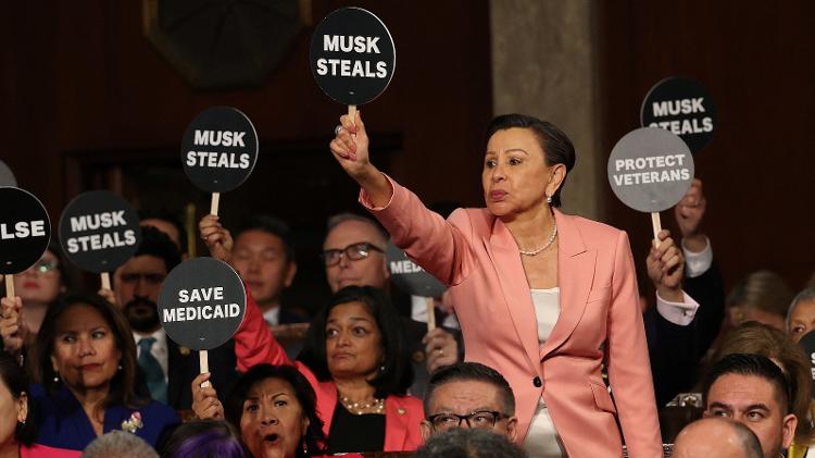 Congressista Nydia Velazquez segura uma placa de protesto com os dizeres "Musk rouba"durante discurso do presidente dos EUA Donald Trump em sessão conjunta no Capitólio