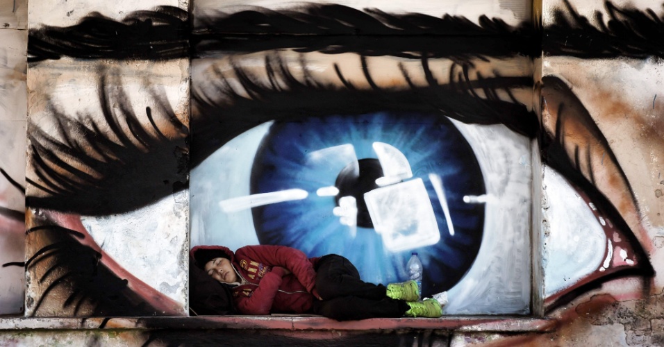06.out.2015 - Mulher refugiada dorme em janela pintada com o grafiti de um olho, no porto de Mytilene, na ilha de Lesbos, Grécia