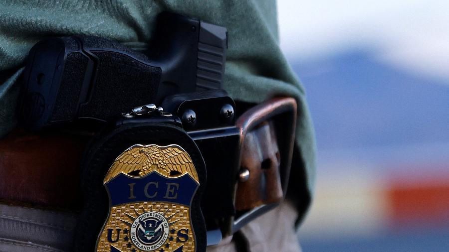 Crachá de um agente do ICE é visto durante uma operação em que migrantes são transferidos para um avião
