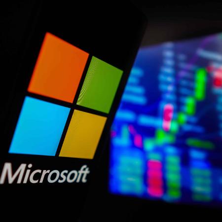 Logo da Microsoft em frente a painel com bolsa de valores Logo da Microsoft em frente a painel com bolsa de valores