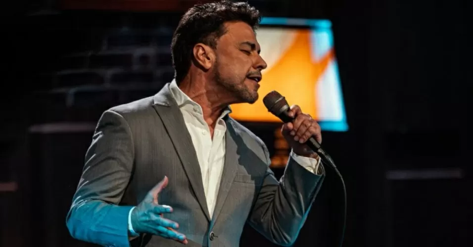 Zezé Di Camargo cantando em microfone, de olhos fechados e roupa social cinza e branca