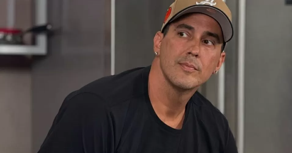 André Marques com camiseta preta e boné marrom, olhando para o lado com expressão desconfiada, inclinado para a frente