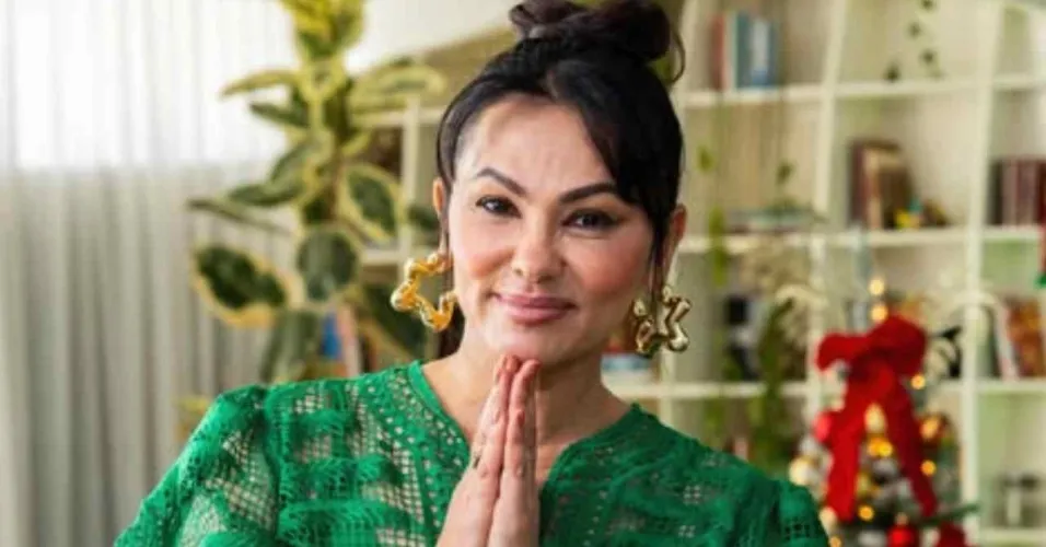 Suzana Alves de verde com mãos juntas e fundo repleto de livros