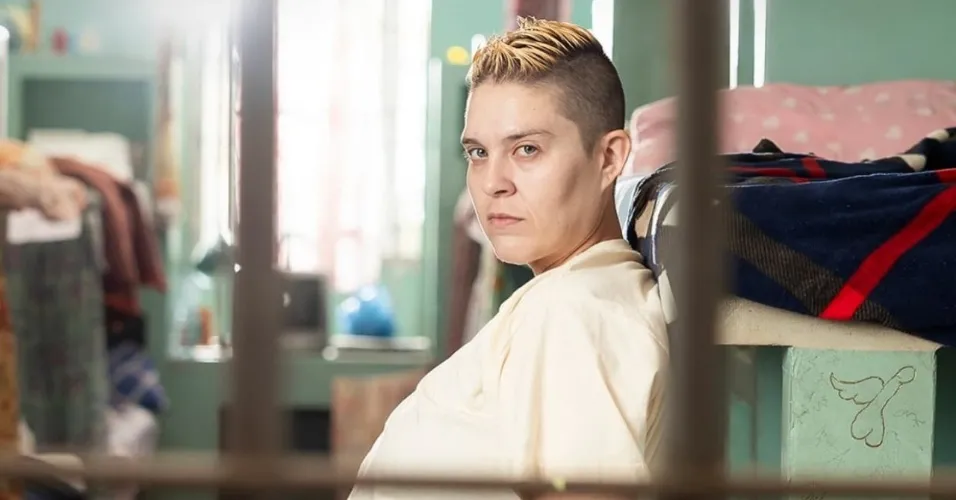 Letícia Rodrigues como a Sandrão de Tremembé, atrás das grades, séria, de camiseta branca larga